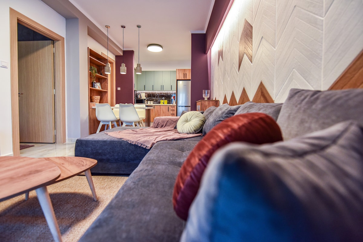 A modern living area features a spacious gray sofa adorned with colorful cushions. Two small wooden tables are positioned in front. Soft lighting highlights the textured wall behind, while an open kitchen is visible in the background, showcasing contemporary cabinetry and appliances.
