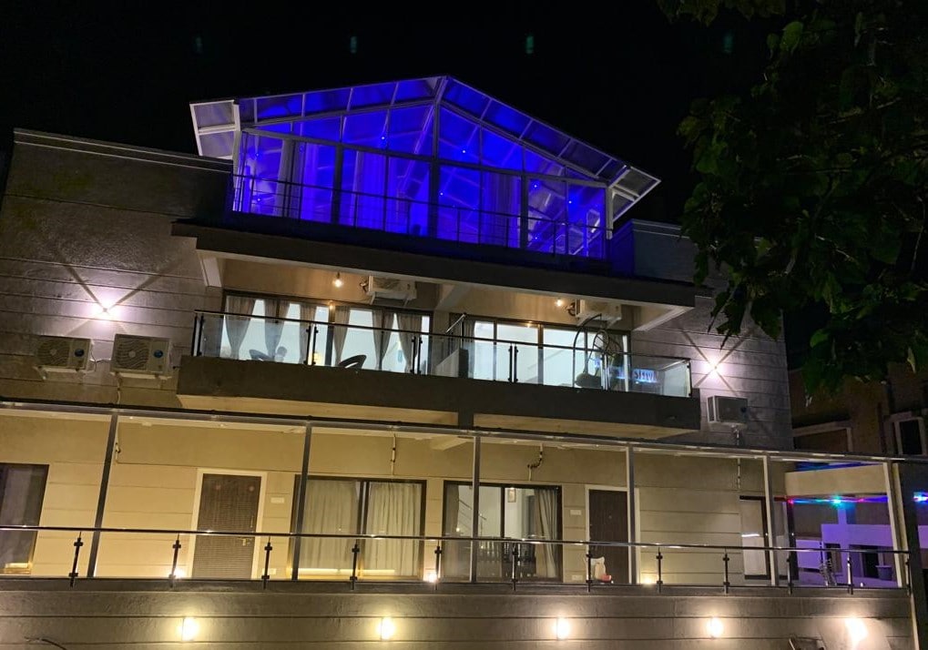 The exterior of the twin bungalow unit is illuminated by soft blue lighting at night. A modern glass house atop the structure features panoramic windows, offering a clear view of the surroundings. A spacious terrace is visible, enhancing the overall aesthetic of the design.