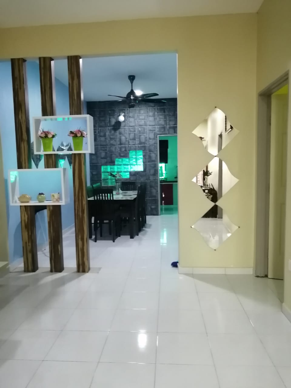 A modern hallway features a glossy tile floor and colorful accent walls. Shelving units with decorative plants are positioned on one side, while a dining area is visible at the end, highlighted by subtle lighting. A mirrored wall decor adds depth to the space.