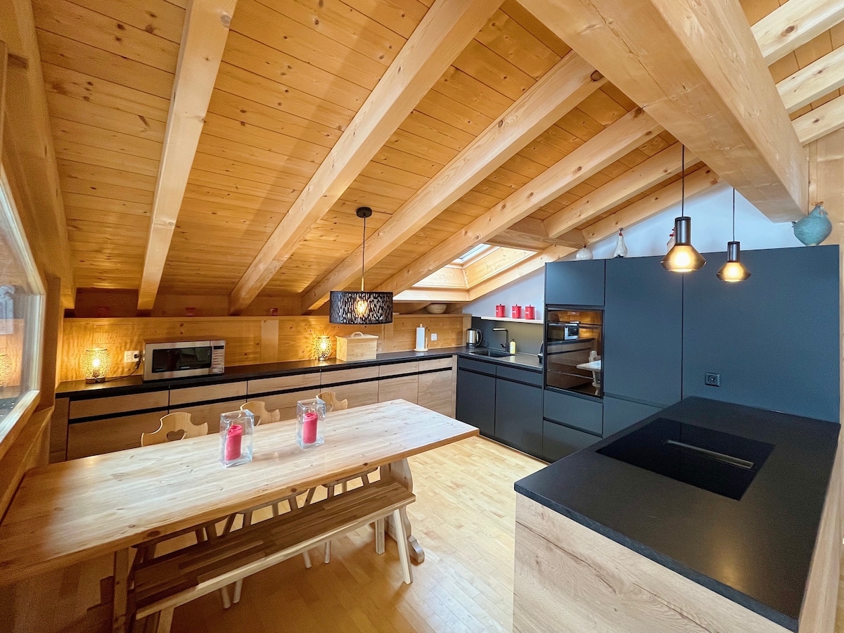 An inviting kitchen space features wooden beams overhead and is dominated by dark cabinetry. A large wooden dining table is positioned centrally, accompanied by light-colored chairs. Modern appliances, including a microwave and oven, are integrated into the design. Natural light is provided through windows.