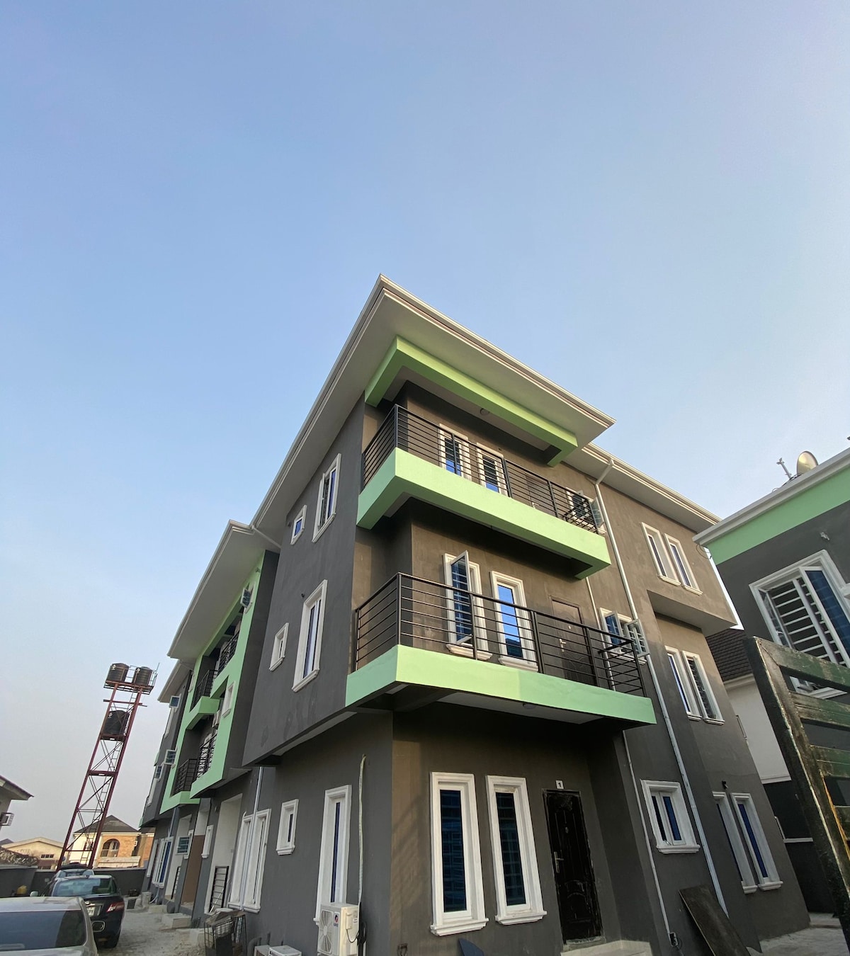 The exterior of a three-story apartment building is captured under a clear blue sky. The structure features a modern design with varying shades of grey and green accents. Multiple balconies with railing enhance the façade, while rows of windows provide natural light to the interior.