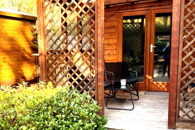 An outdoor seating area features a pair of black chairs positioned around a small table, set against a backdrop of wooden lattice. Lush greenery surrounds the space, while glass doors provide access to the interior of the vacation home.