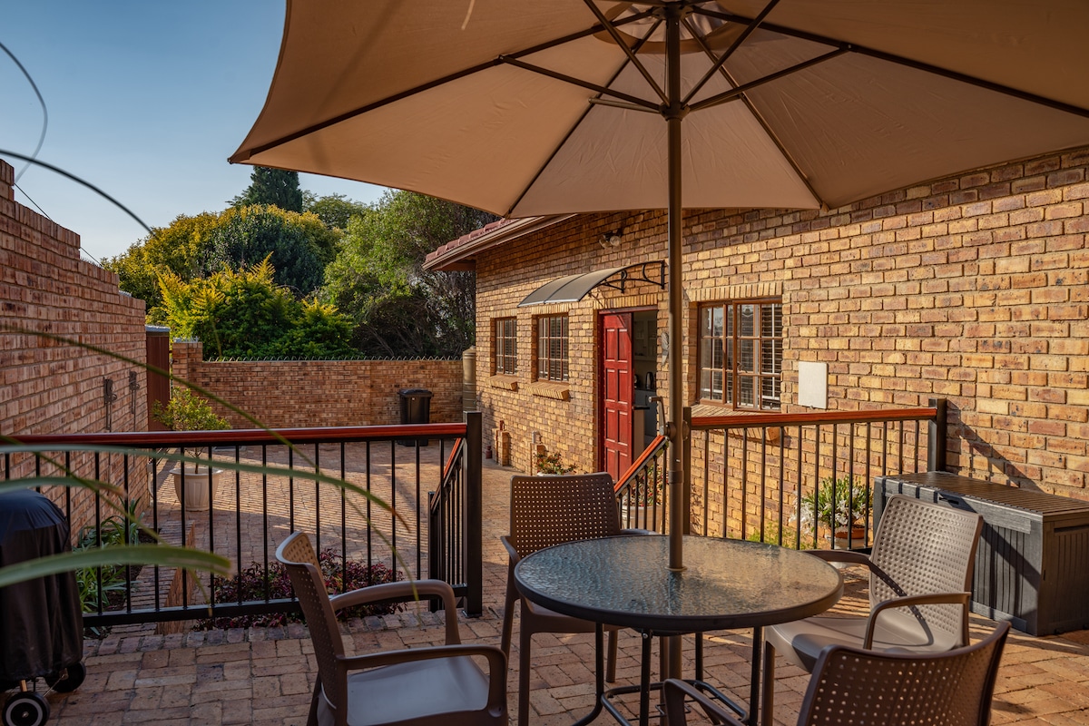 An outdoor patio area is shaded by a large umbrella over a round glass table surrounded by four chairs. The brick walls create a cozy atmosphere, with a glimpse of greenery and a red door visible in the background.