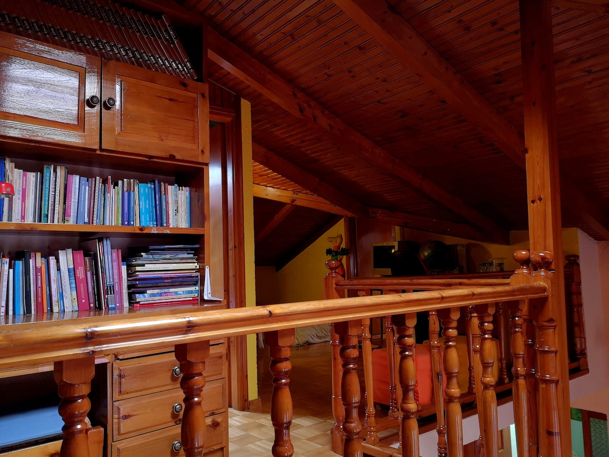 An airy loft space is visible, featuring wooden beams and a warm-toned ceiling. Bookshelves filled with various titles are positioned against one wall. A polished wooden railing provides a view into the area beyond, while soft lighting enhances the inviting atmosphere.