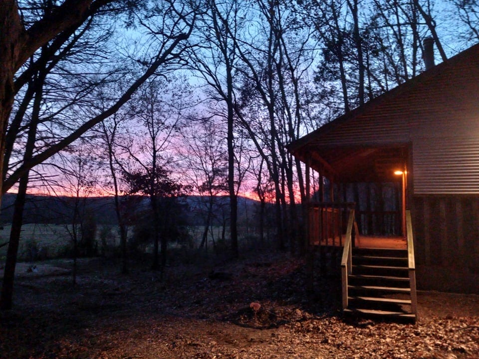 The exterior of a two-story cabin is captured at dusk, with a warm glow from a porch light illuminating the entrance. Trees frame the image, while a colorful sunset creates a serene backdrop over the hillside in the distance.