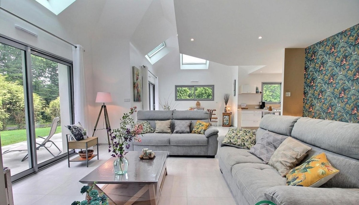A spacious living area features large glass doors that invite natural light, with views of the garden. Two comfortable sofas are arranged around a simple coffee table, complemented by decorative cushions. A modern kitchen can be glimpsed in the background, enhancing the open layout.