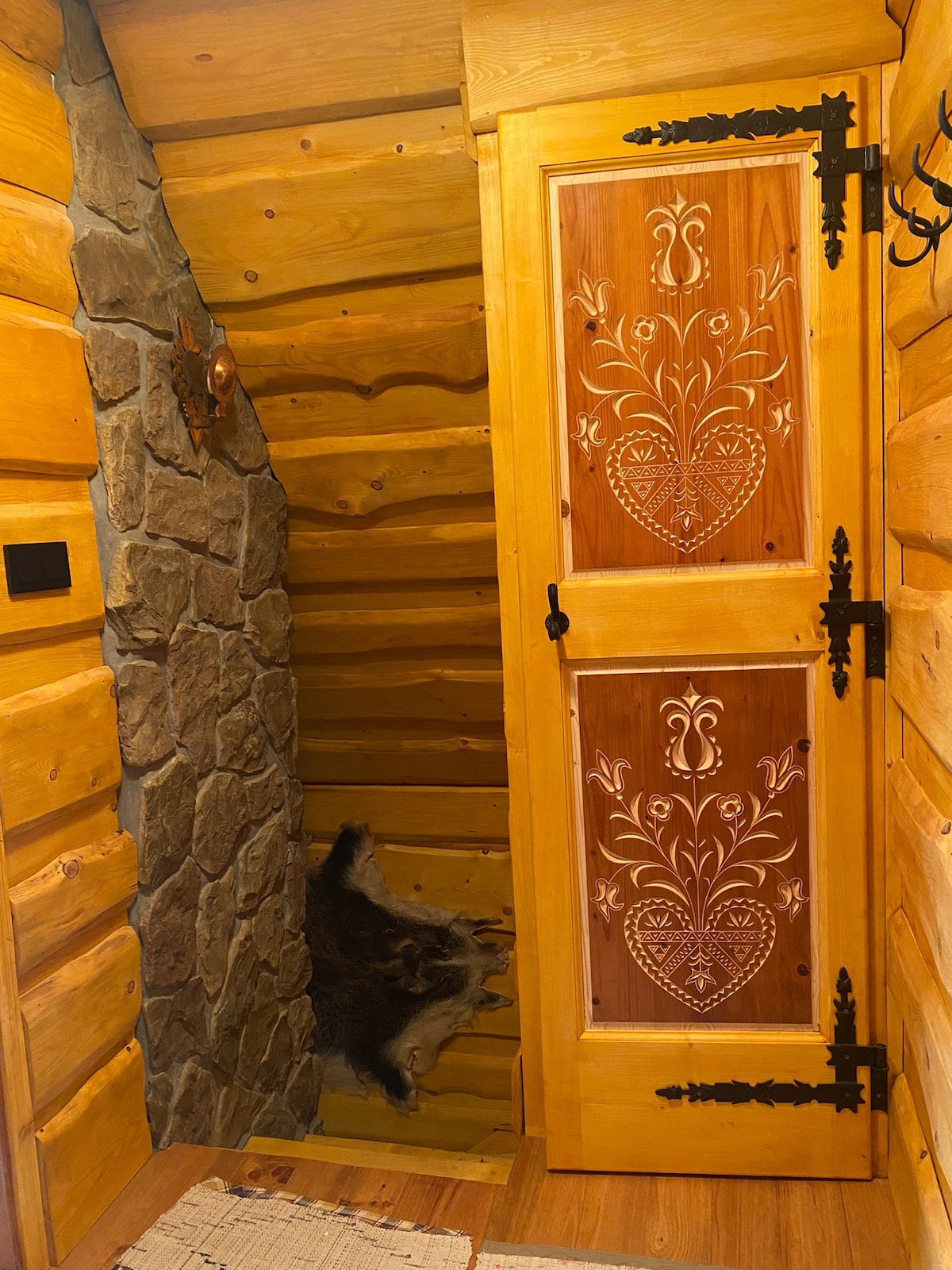 A wooden door with intricate carvings is displayed, leading to a staircase. The walls are made of natural wood, complemented by a stone feature wall. A decorative rug lies on the wooden floor, enhancing the cozy entryway.