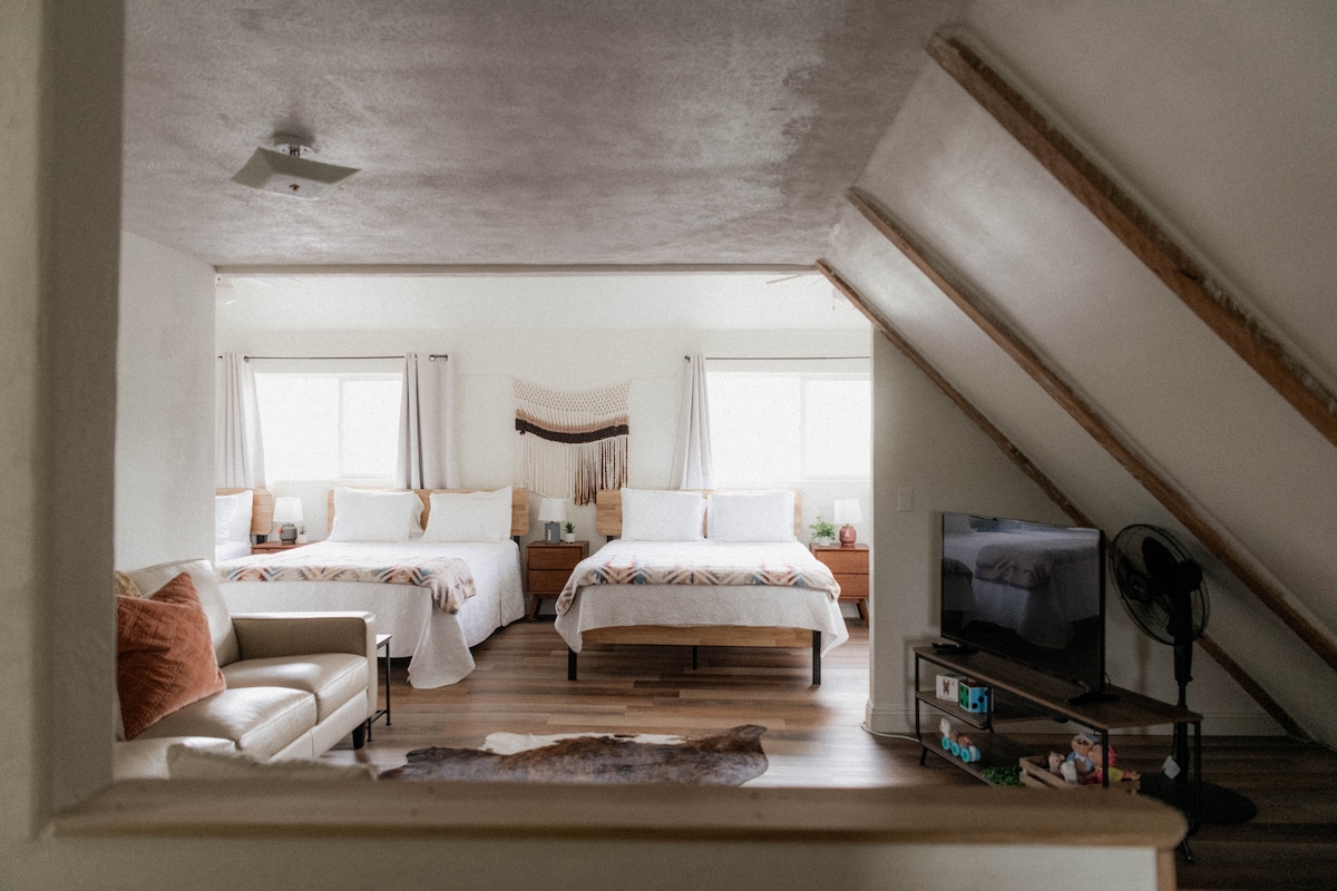 An open loft area is visible, featuring four queen beds with decorative throw blankets and bedside tables. Natural light streams in through two windows, illuminating the wooden floor. A cozy seating arrangement includes a light-colored sofa, while a television is positioned nearby.