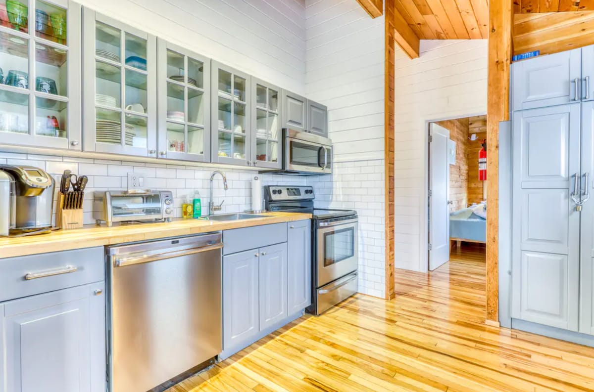 The kitchen features modern stainless steel appliances, including a stove and dishwasher. Ample cabinetry displays dishware and glassware, while hardwood floors add warmth to the space. A doorway leads to the adjacent living area, enhancing the open concept layout.