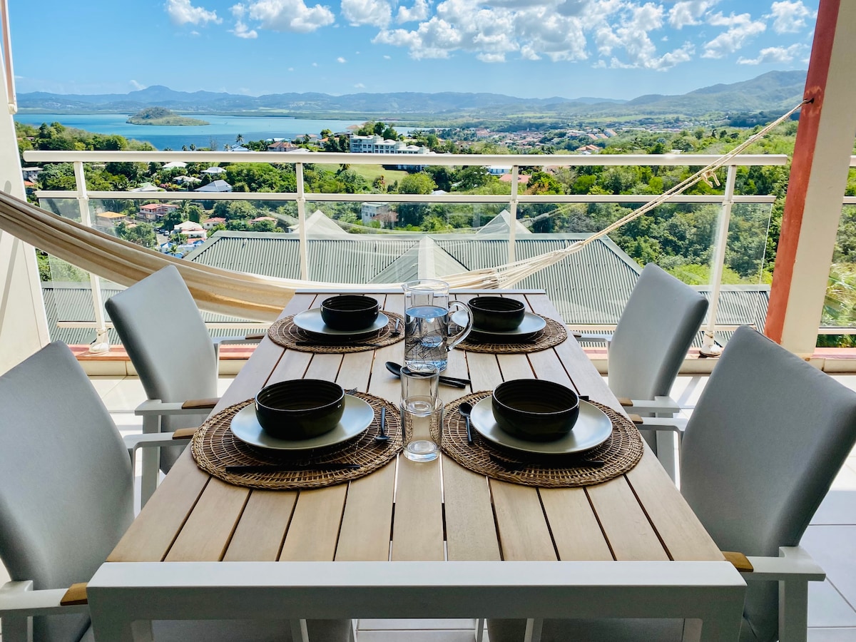 A dining table set for four is positioned on a terrace, overlooking a scenic view of the sea and hills. Each place setting includes a plate, bowl, and glass. A hammock can be seen in the background, enhancing the outdoor relaxation space.
