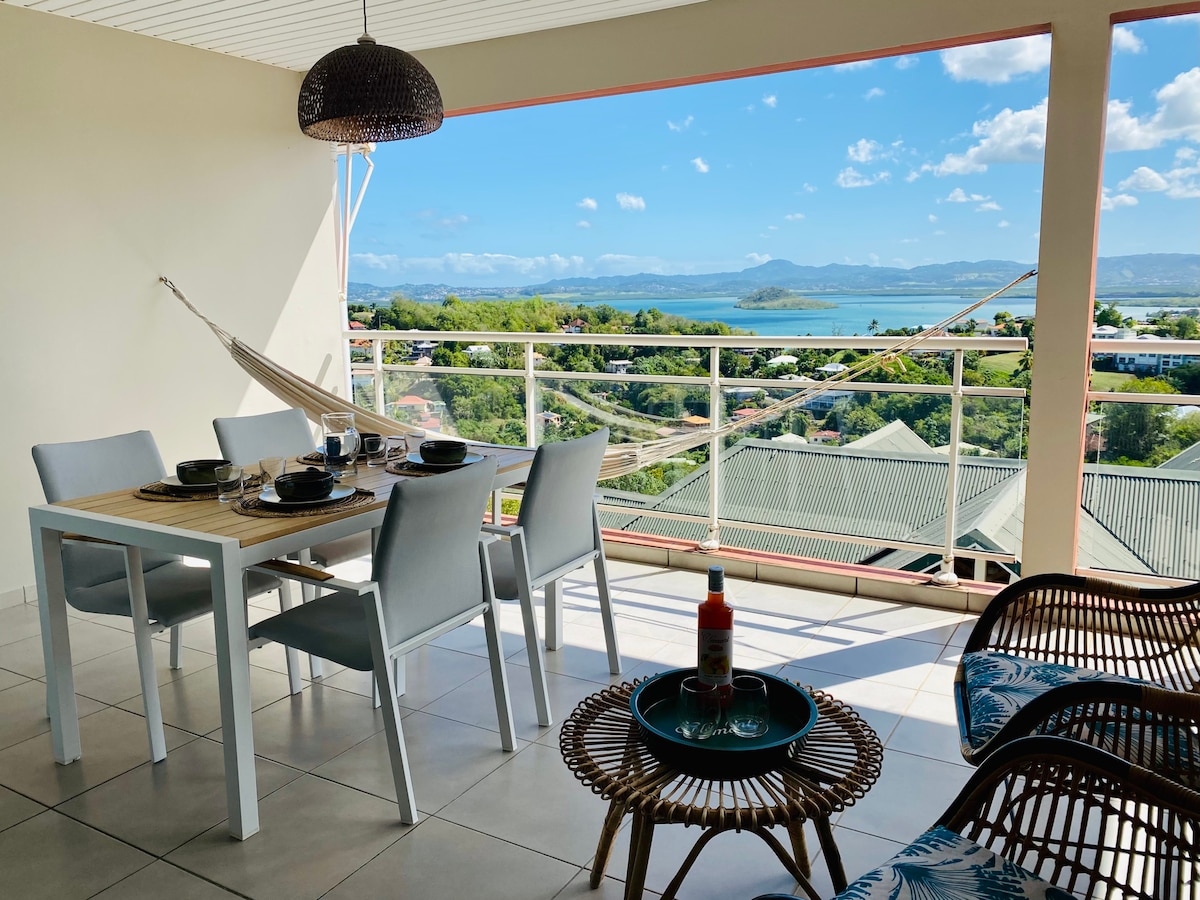 An outdoor terrace features a dining table set for four with plates and glasses arranged neatly. A hammock hangs nearby, providing a relaxing spot. Comfortable seating areas with wicker chairs integrate with the spacious terrace, while views of the sea and surrounding landscape can be seen in the background.