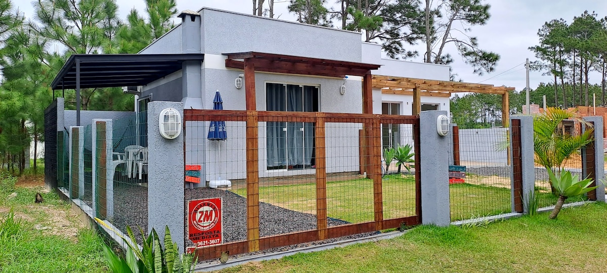 A private bungalow is surrounded by a lush green yard with a fenced area. White outdoor furniture is visible beneath a shaded awning. The exterior features a modern design with large windows and a welcoming entrance, framed by trees in the background.