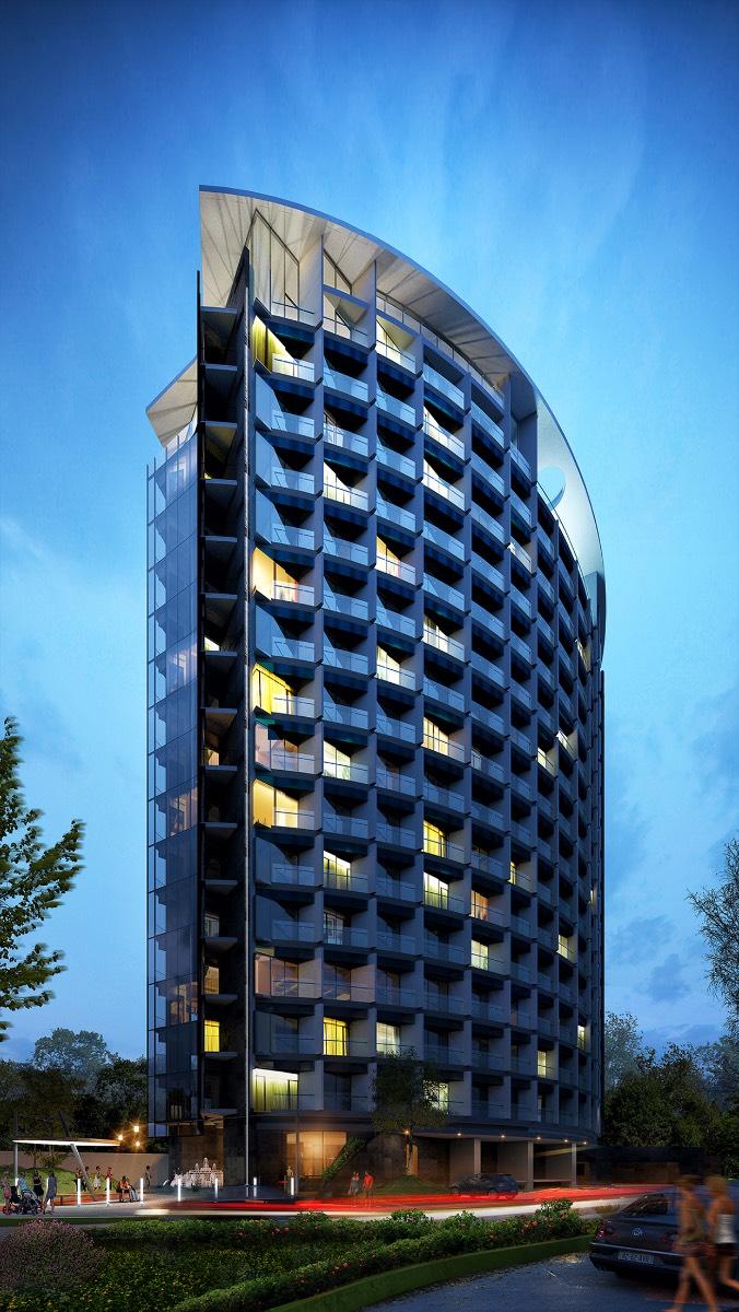 The modern building features a unique curved design, showcasing multiple balconies framed by large glass windows. Softly illuminated by exterior lights, the structure stands against a twilight sky, surrounded by landscaped greenery and pathways leading to the entrance.