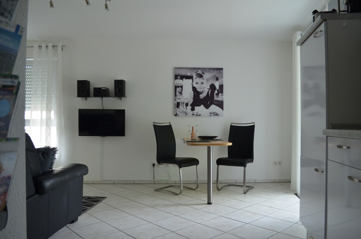 A cozy seating area is presented with a small round table and two black chairs. A sleek black sofa is positioned nearby, while a wall-mounted television and speakers add modernity. A framed black-and-white image decorates the wall, adding a touch of character to the space.