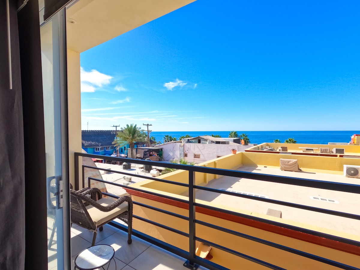 A private balcony offers expansive views of the ocean and surrounding rooftops. A small round table and a wicker chair are positioned for relaxation, while bright blue skies enhance the inviting atmosphere of the outdoor space.