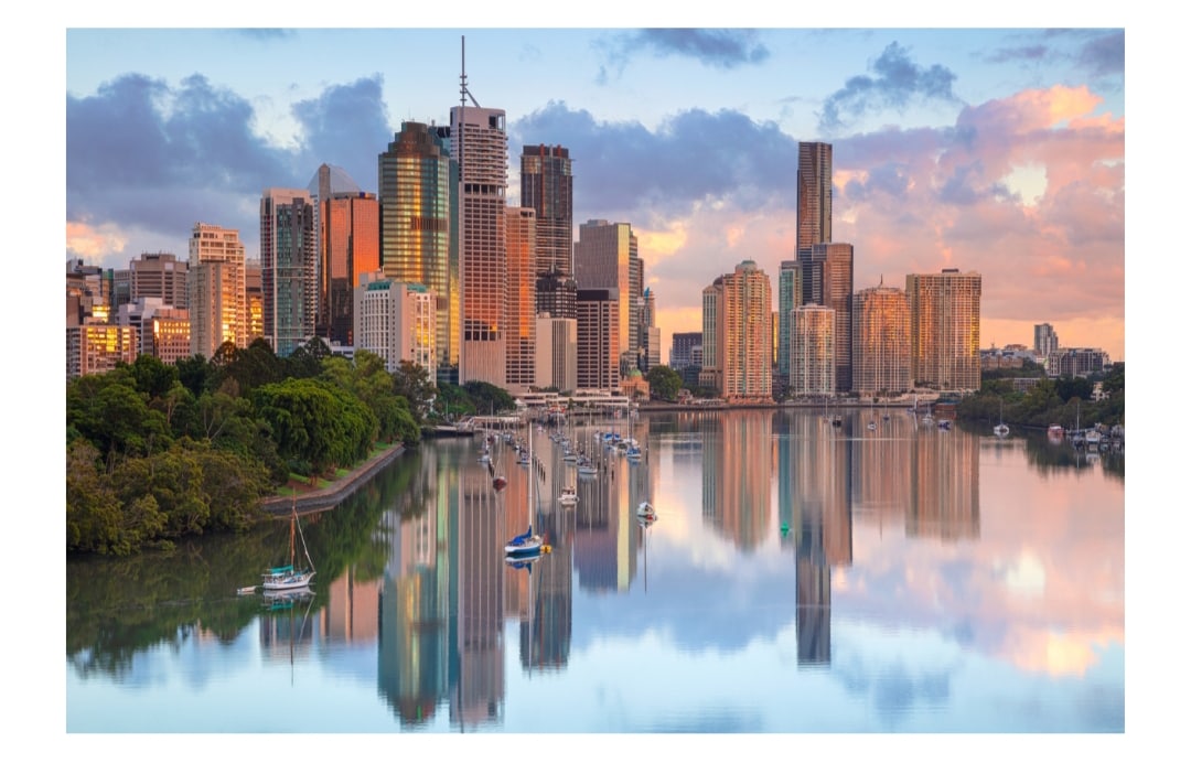 The city skyline is reflected in serene waters, showcasing modern high-rise buildings adjacent to lush greenery. Soft hues of sunrise illuminate the scene, with boats moored along the water's edge, creating a tranquil urban landscape.