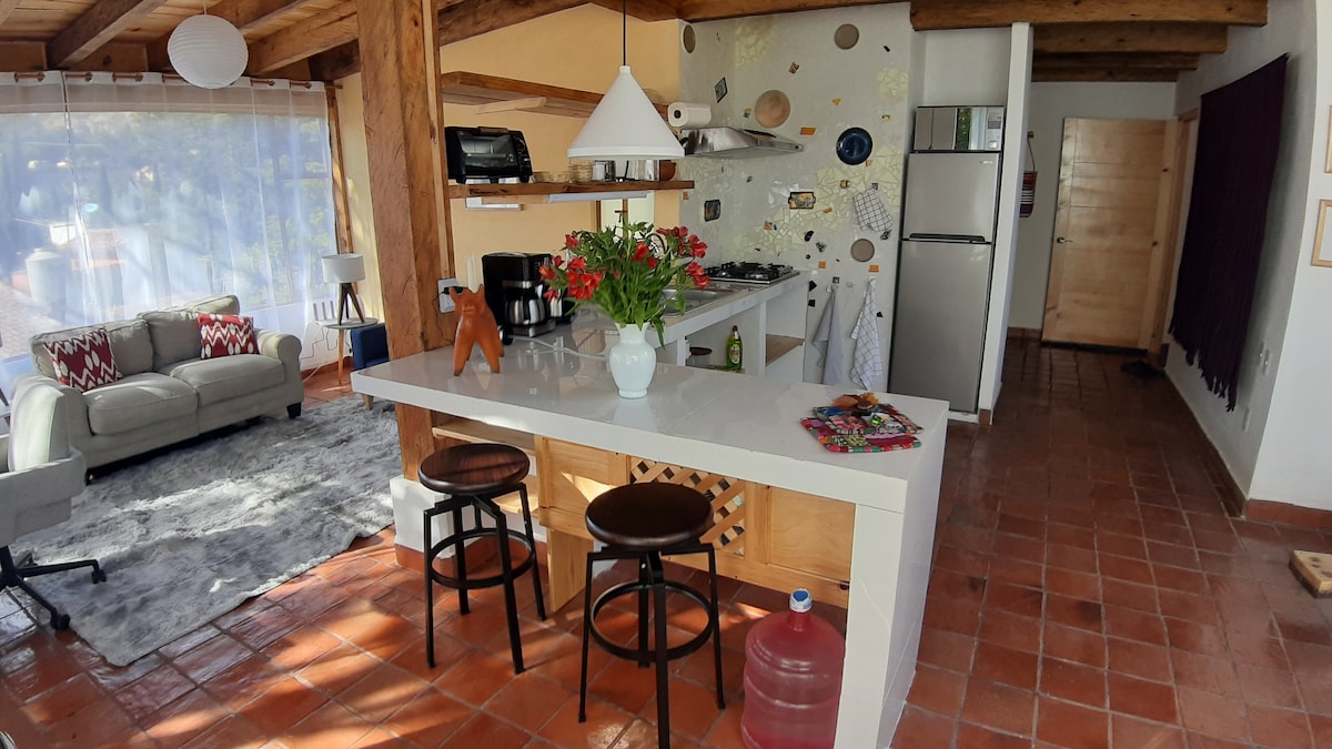 An open kitchen area is visible, featuring a modern design with a breakfast bar and two stools. A vase of fresh flowers decorates the counter, while a cozy seating area with a light-colored sofa is positioned nearby. Warm wood beams and terracotta tiles add natural texture.