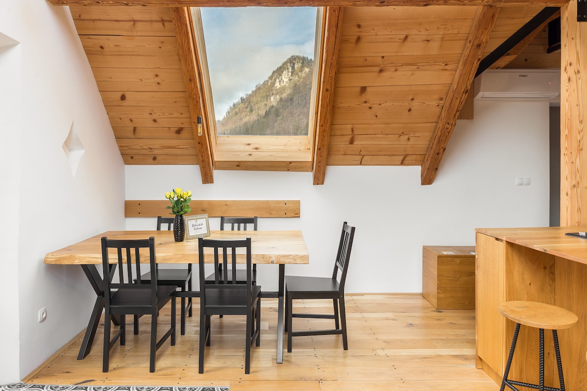 An inviting dining area showcases a wooden table surrounded by four black chairs. A small bouquet of yellow flowers decorates the table. Two skylights above provide natural light, offering a view of the surrounding hills. Warm wooden beams are featured in the ceiling.