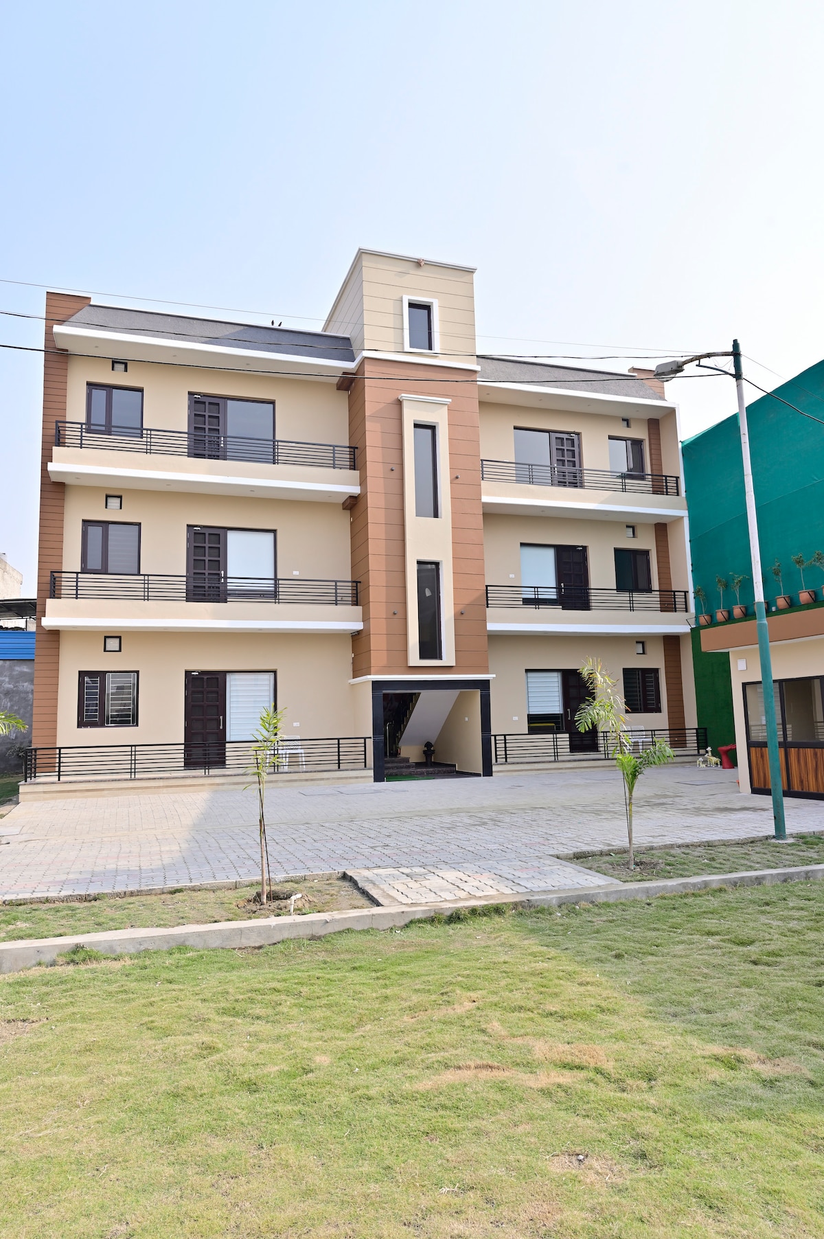 The three-story building displays a modern architectural design featuring multiple balconies and large windows. A manicured lawn stretches in front, accompanied by a paved pathway and young trees lining the entrance. The exterior is complemented by a blend of neutral and warm tones.