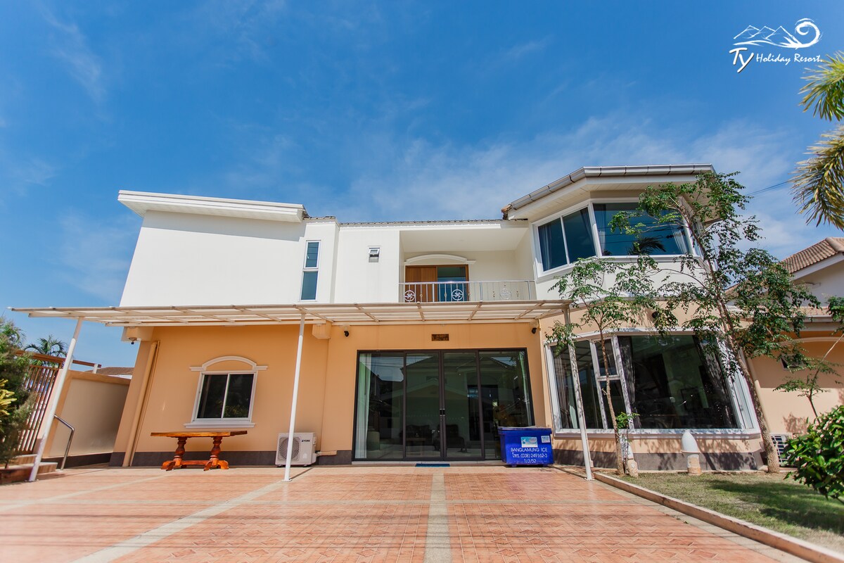The exterior of the property features a spacious entrance with large glass doors. A covered patio area is highlighted, alongside a wooden table. Lush greenery is present, adding to the inviting outdoor atmosphere. The building boasts a combination of modern architectural elements.