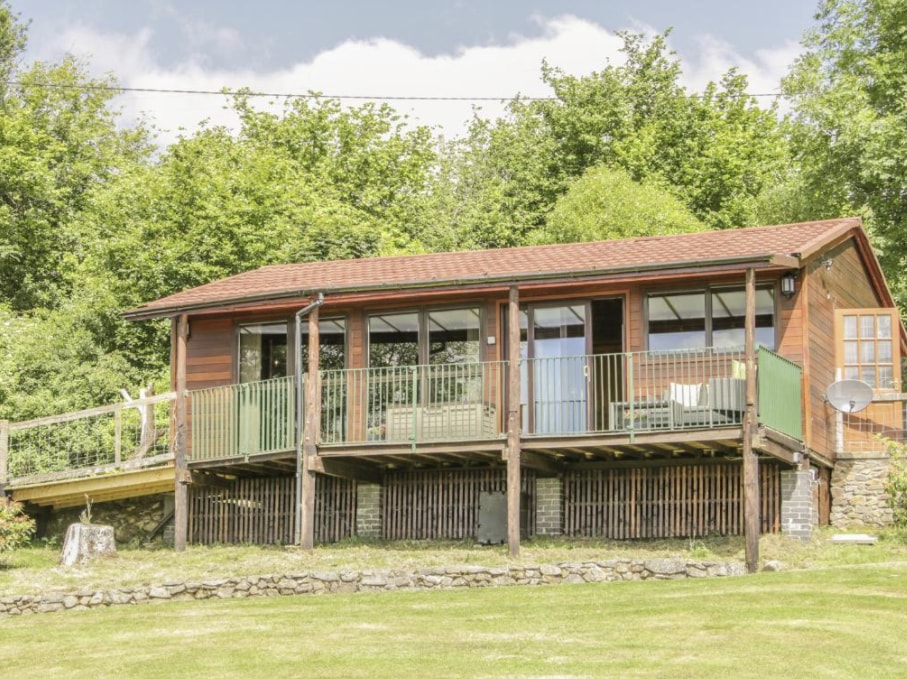 The cabin features a wooden exterior with large glass windows and a wraparound balcony. Surrounding greenery provides a natural backdrop, while a stone wall lines the base. The elevated structure allows for views of the landscape below.