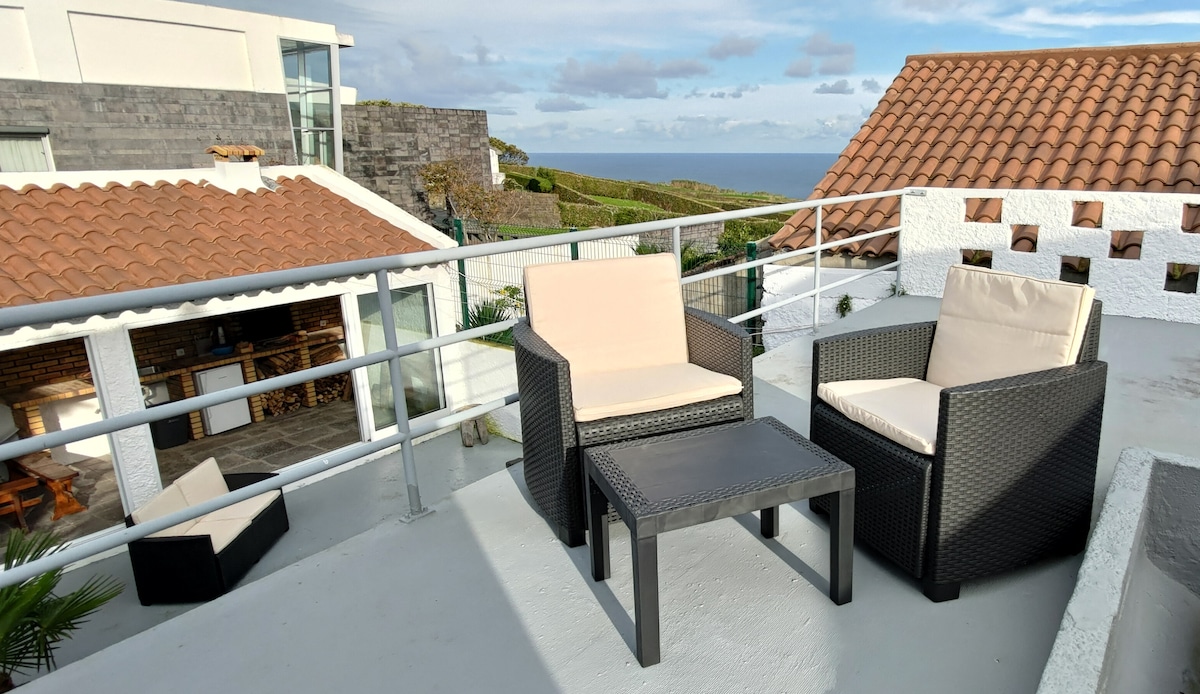 A cozy terrace features two wicker chairs and a small table, providing a relaxing space with views of the ocean and surrounding landscape. The area is illuminated by natural light, highlighting the modern architecture and the traditional tiled roofs in the background.