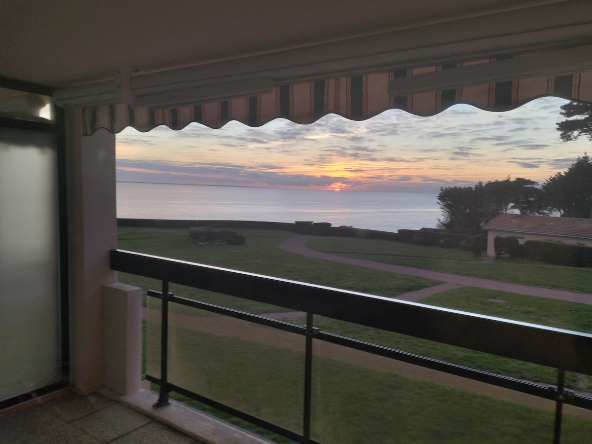 A spacious balcony offers a serene view of the sea at sunset, with warm hues reflecting on the water. A grassy area is visible below, bordered by greenery. The protective awning is partially open, framing the tranquil scene.