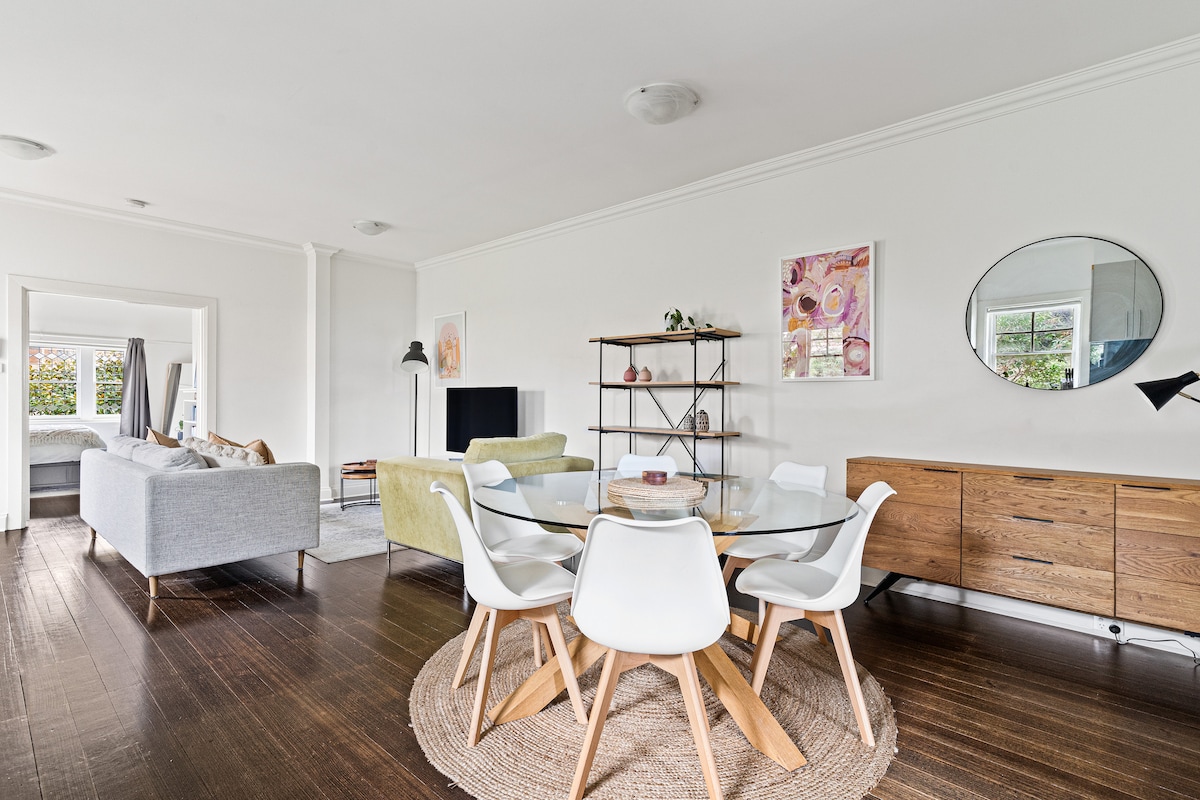 An open plan living and kitchen area is featured with hardwood flooring, natural light entering through the windows. A round dining table with white chairs occupies the center, while a green sofa and grey seating are arranged nearby for relaxation. Decorative accents enhance the modern aesthetic.