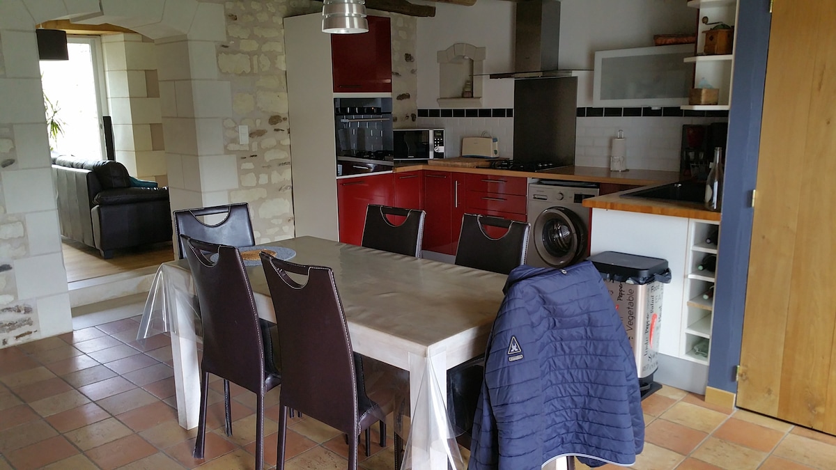 A spacious dining area is highlighted by a rectangular table surrounded by brown, high-backed chairs. The kitchen features red cabinets and modern appliances, with a view of the living area beyond. Natural light enters through large windows, accentuating the tile flooring.