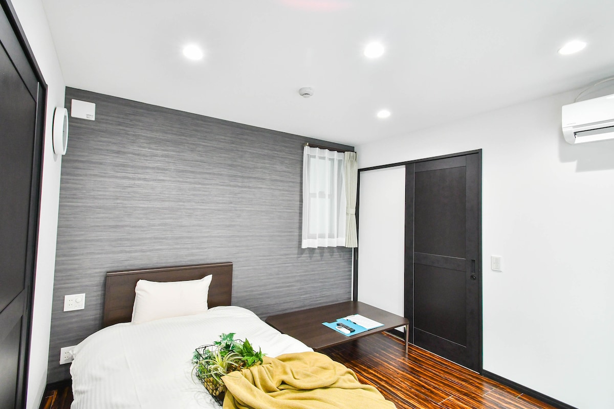 A modern, minimalist room features a spacious bed dressed in white linens, complemented by a cozy blanket. A simple wooden table is included, along with a window dressed in sheer curtains. The walls display a textured grey finish, and overhead lights provide bright illumination.