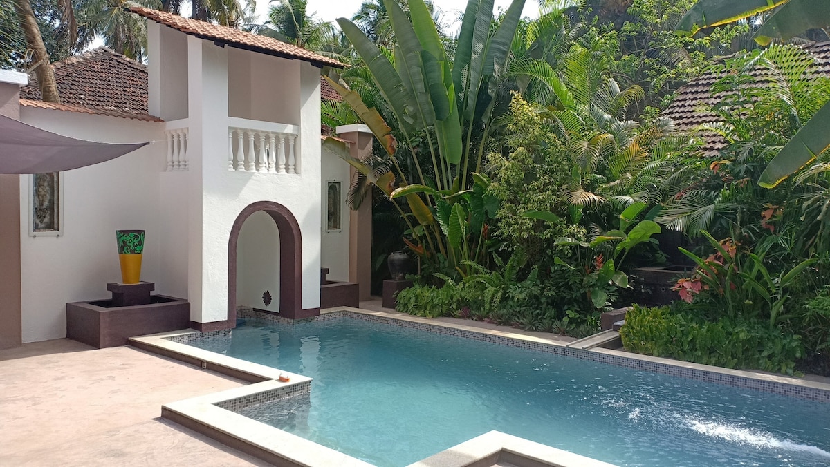 A serene outdoor area features a private pool surrounded by lush tropical plants and vibrant flowers. A distinctive archway leads to a separate structure, adding architectural interest. Sunlight filters through the leaves, creating a tranquil atmosphere for relaxation.