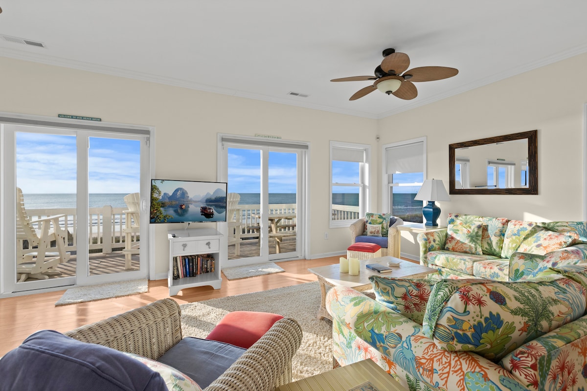 A bright living room offers panoramic ocean views through large glass doors. Colorful seating surrounds a coffee table, and a television is positioned on a stand. Light wood flooring complements the coastal decor, creating an inviting space for relaxation.