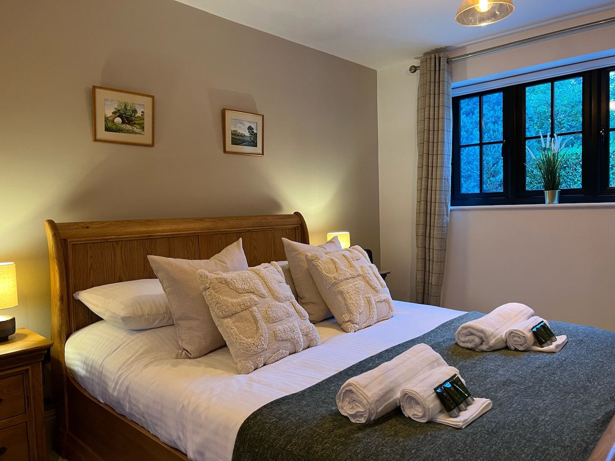 A spacious sleeping area features a wooden bed with soft linens, accompanied by two decorative pillows. Nightstands with lamps are situated on both sides. A window with a plaid curtain allows natural light, highlighting the serene wall decor, which includes two framed images.