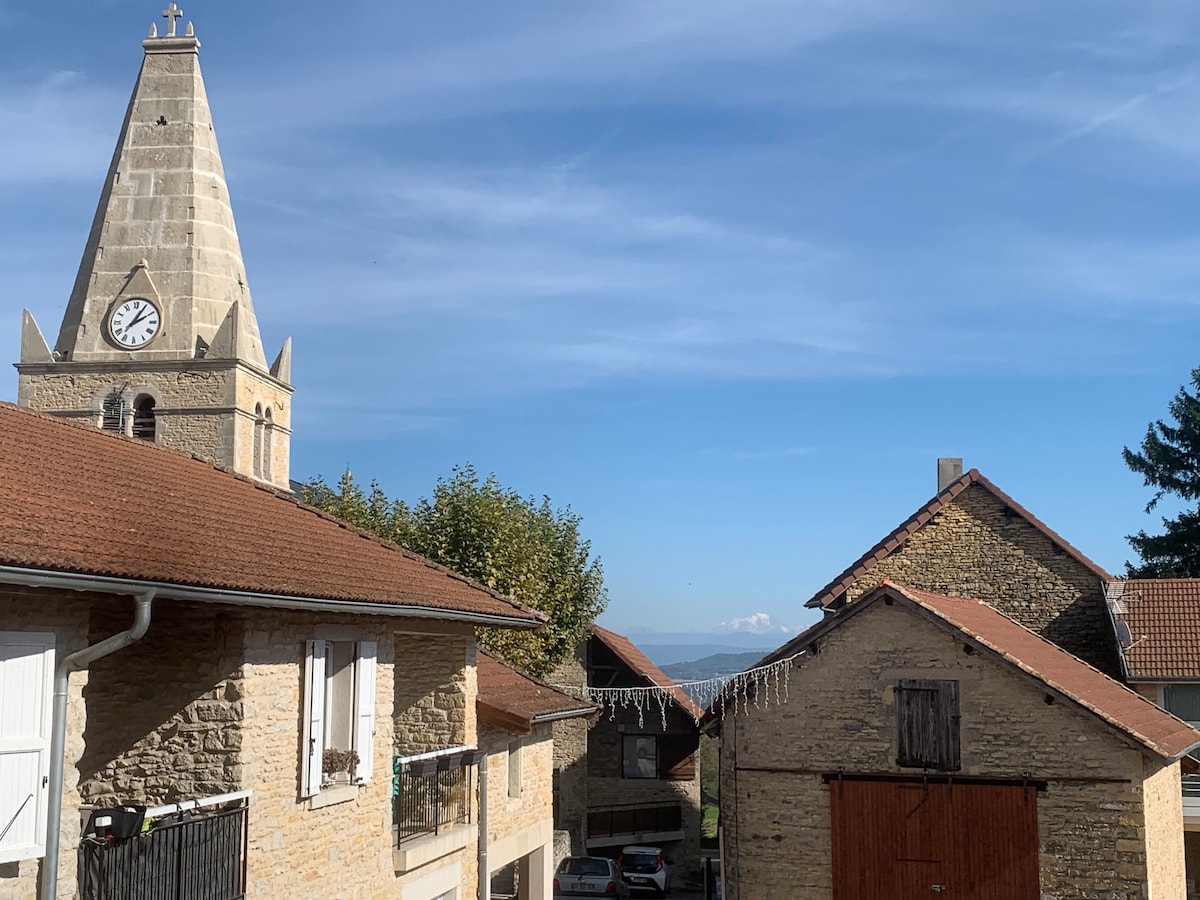 The image showcases a quaint village scene, featuring a stone house with a balcony, a clock tower with a peaked roof, and a rustic barn. Clear blue skies provide a backdrop, while distant hills are visible in the background.