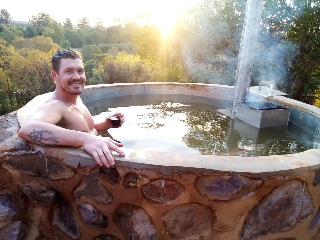 A hot tub is set on a stone platform, surrounded by greenery. Steam rises softly from the water, reflecting the warm light of the setting sun. The serene atmosphere offers a tranquil space for relaxation amidst nature.
