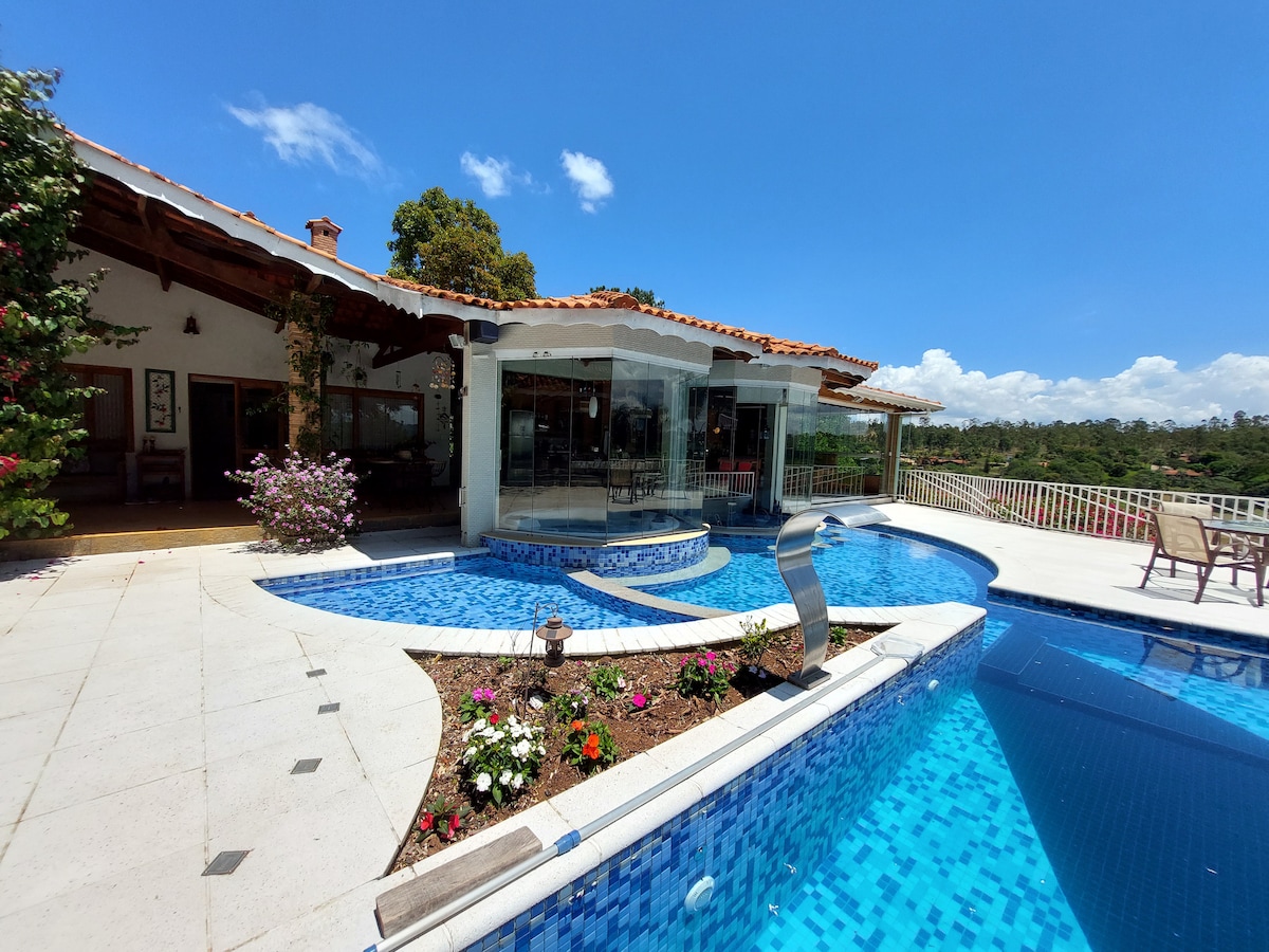 A spacious outdoor area features a large swimming pool surrounded by colorful flower beds. The house, with large glass windows, provides a seamless connection to the outdoors. Sunlight brightens the scene, highlighting the inviting blue tiles of the pool and the greenery in the background.