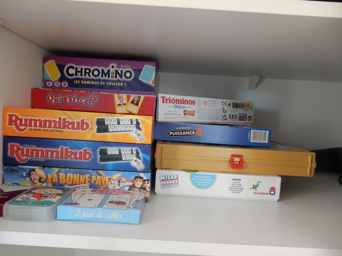 Several board games are neatly arranged on a shelf. Boxes of Rummikub, Chromino, and Quizzace are prominently displayed, alongside a Triominos box and a yellow game case. Various card games are also visible in the foreground.