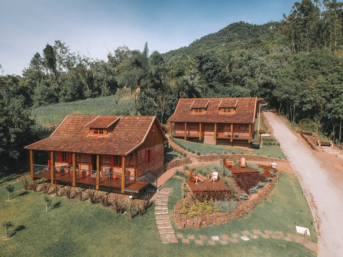 Two colonial-style chalets are positioned in a green landscape, with wooden exteriors and sloped roofs. Paths lead to the entrances, with lounge chairs arranged on the lawn. Lush vegetation frames the chalets, complemented by distant hills in the background.