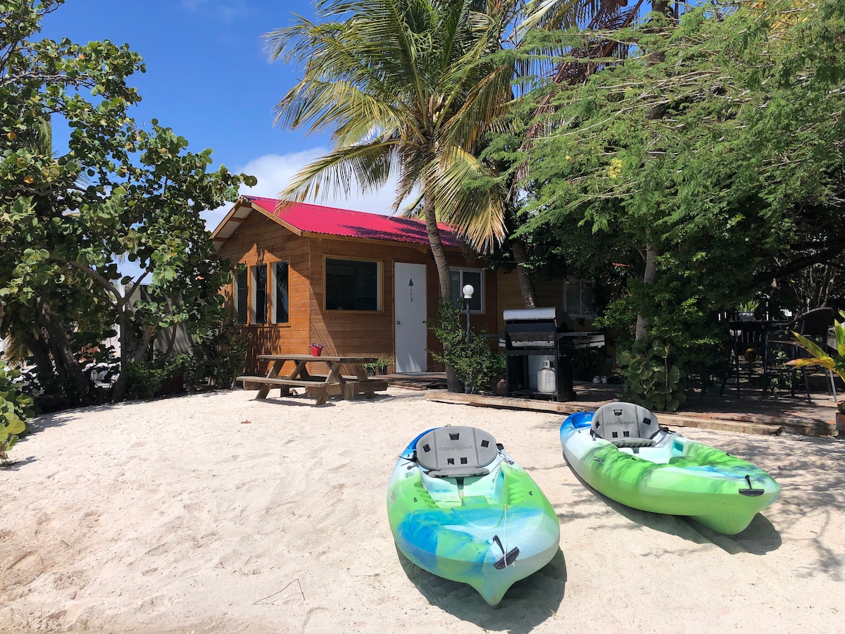 A charming wooden cabin is surrounded by sandy terrain and tropical greenery. Two kayaks are positioned in front, with a picnic table nearby. A grill stands to the side, emphasizing the outdoor space available for guests to enjoy.