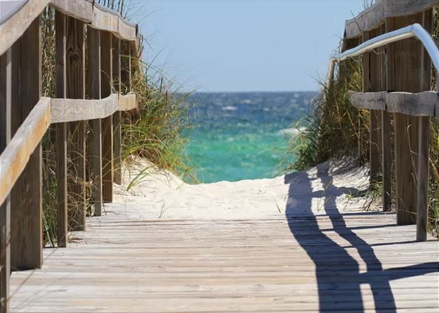 A wooden walkway is framed by tall beach grass, leading toward the tranquil ocean. The sandy path is bordered by wooden railings, and the inviting turquoise waters are visible in the distance under a clear blue sky.
