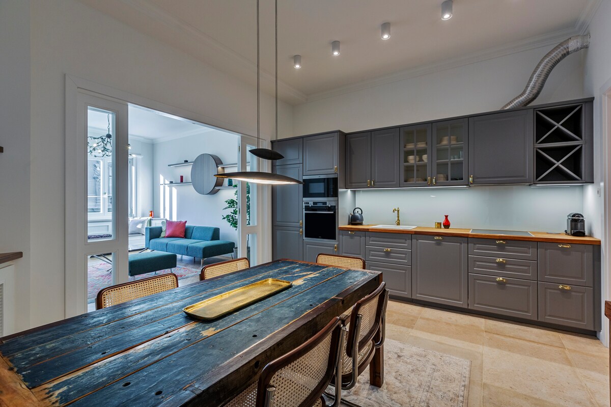 A spacious dining area features a rustic wooden table surrounded by six rattan chairs, under a contemporary pendant light. A well-equipped kitchen showcases elegant grey cabinetry, modern appliances, and soft backlighting, while a glimpse of a cozy living room is visible through the open doorway.