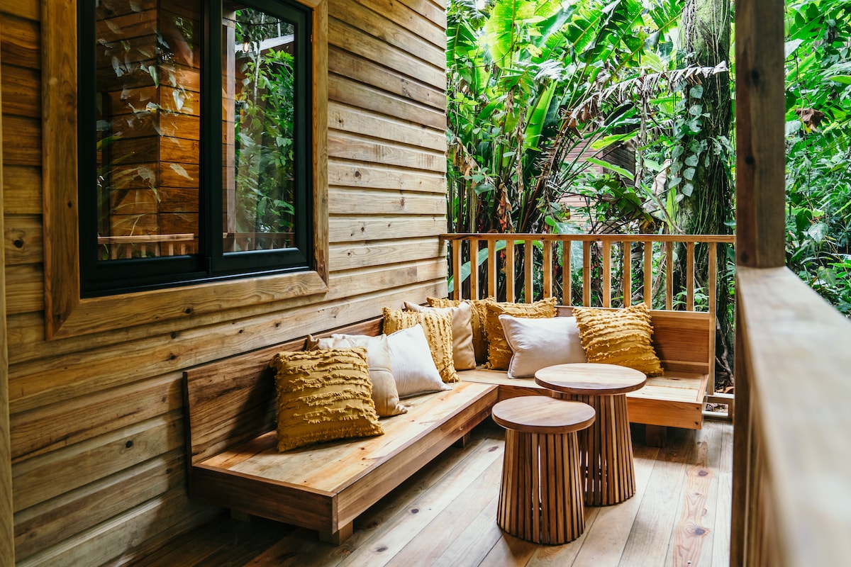 A wooden porch area features a cozy seating arrangement with textured cushions and two circular wooden tables. Lush greenery surrounds the space, creating a serene connection to nature, while large windows allow natural light to illuminate the seating.
