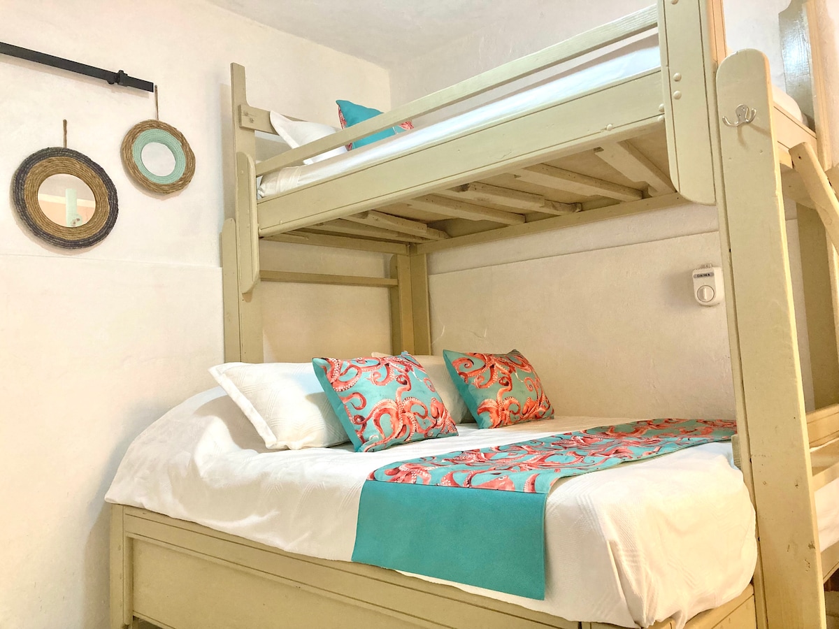 A compact sleeping area features a bunk bed with a spacious lower level. The bed is adorned with decorative pillows in vibrant colors, and a bright turquoise blanket complements the bedding. Circular wall mirrors add a touch of decor to the white walls.