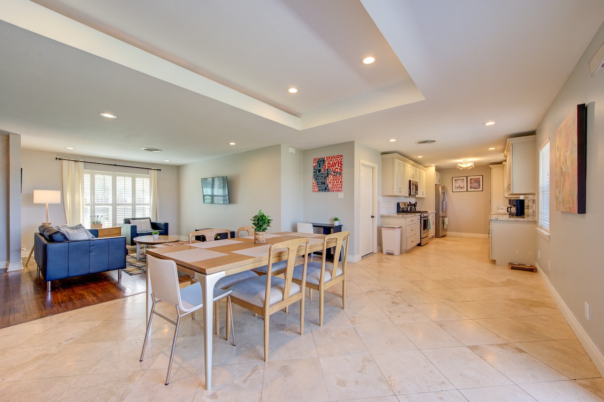 An open living area is showcased, featuring a large dining table surrounded by light wood chairs. The kitchen is visible with modern appliances, while a comfortable seating arrangement is positioned nearby. Natural light fills the space, highlighting the tiled flooring throughout.