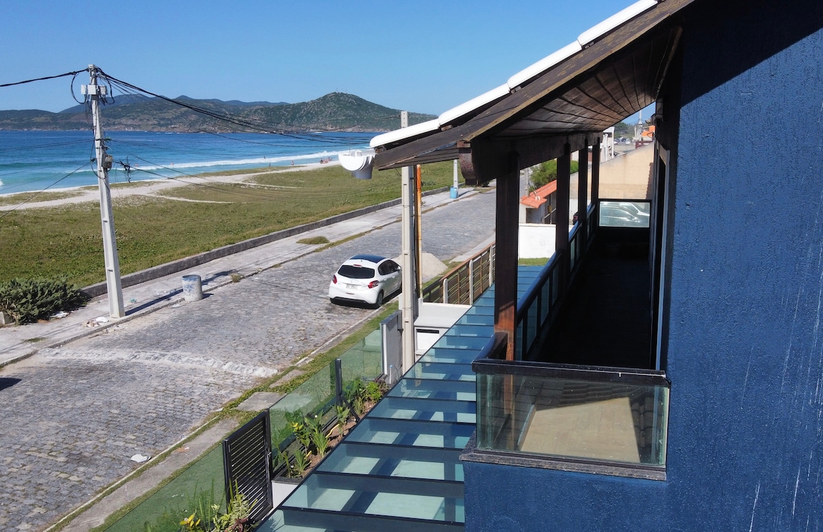 A view overlooks the beachfront from the loft's balcony, with clear line of sight to the sandy shore. A quiet street is visible below, featuring parked car and pathways that lead towards the beach. Natural surroundings complement the coastal scenery.