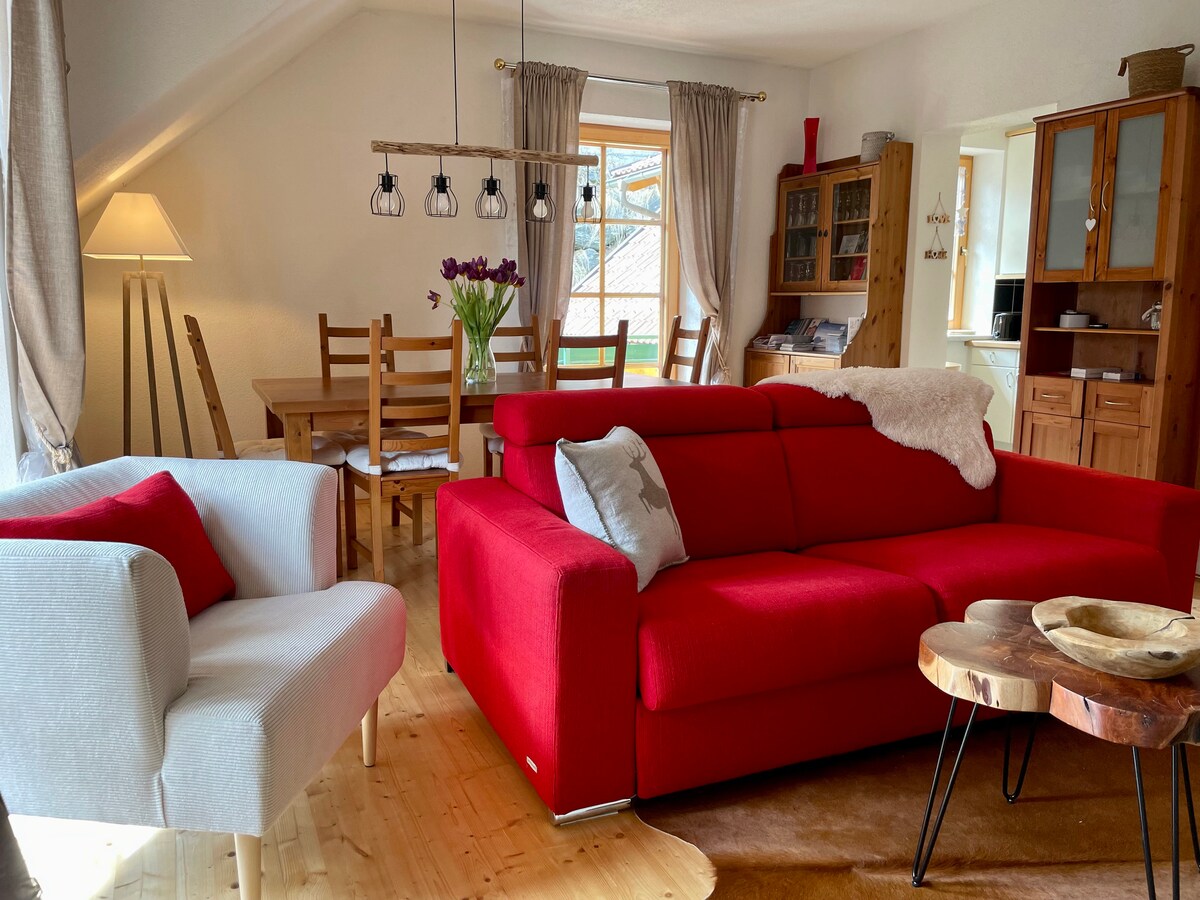 An inviting living space showcases a red sectional sofa accompanied by a white armchair. A dining table and chairs are arranged nearby, illuminated by natural light from large windows. Decorative features include a wooden coffee table and hanging light fixtures, enhancing the cozy atmosphere.