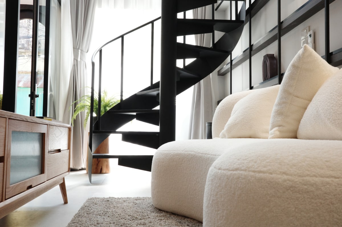 A modern seating area features a spacious, textured couch in a light tone, complemented by a soft rug. A striking spiral staircase in black metal leads to the mezzanine. Large windows allow for ample natural light, framed by sheer curtains.