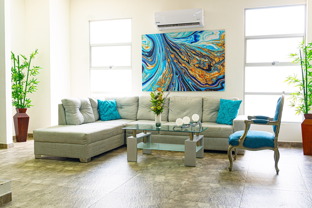 A spacious living area features a light-colored sectional sofa complemented by blue decorative pillows. A glass coffee table sits at the center, adorned with a vase of flowers. The room is brightly lit by natural light from large windows, with greenery in decorative pots enhancing the ambiance.