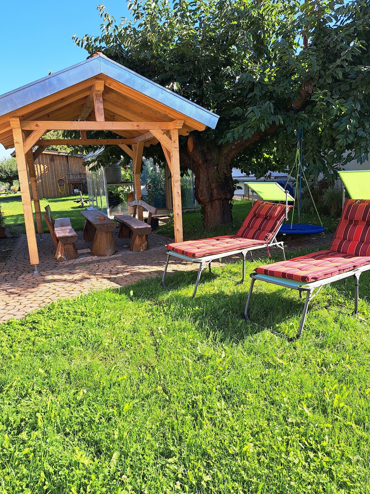 An outdoor space features a wooden gazebo with a rustic table and benches made from logs. Two lounge chairs with red cushions are positioned on the green grass, inviting relaxation. A tree provides shade, and a swing hangs nearby, contributing to a family-friendly environment.