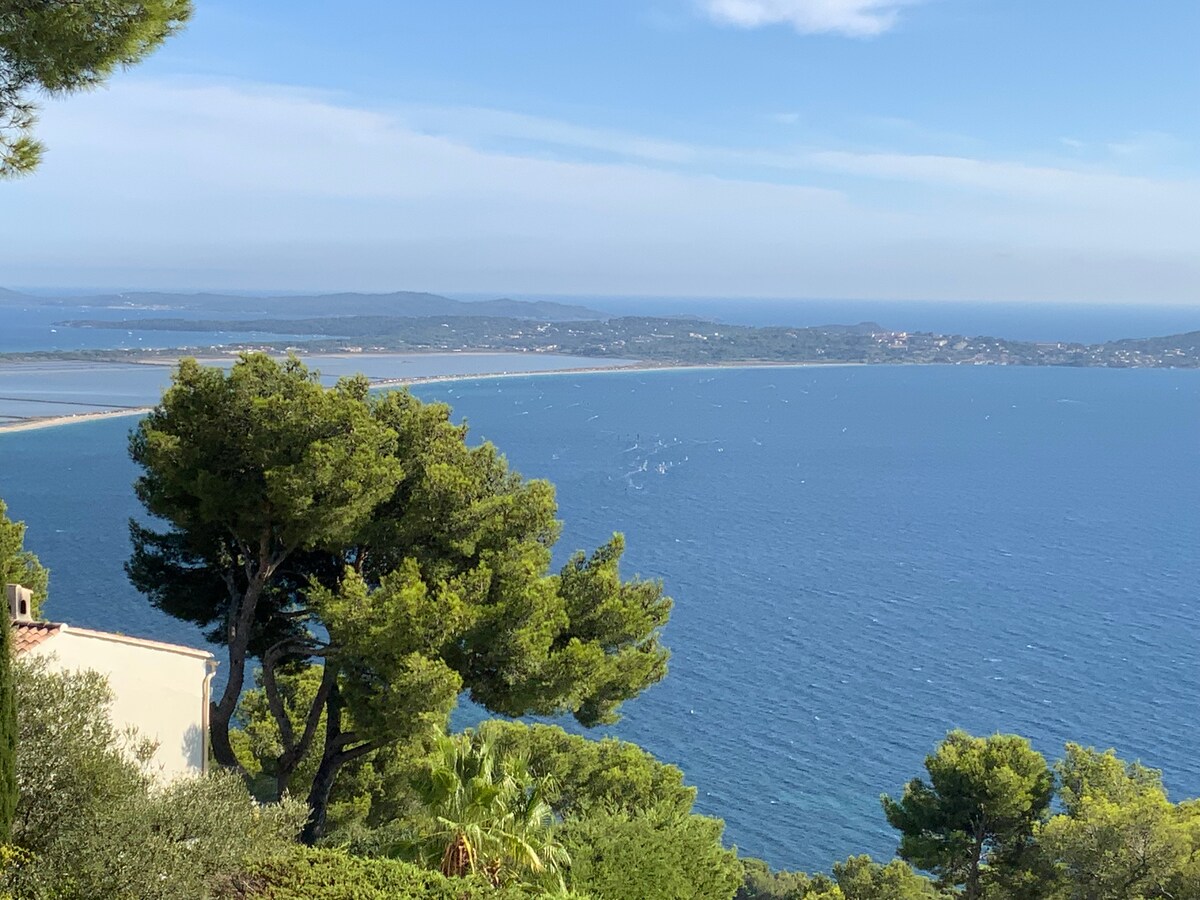A panoramic view showcases the sparkling blue sea, dotted with small boats. Lush green trees frame the scene, leading towards the coastline with sandy shores. The distant islands can be seen on the horizon, enhancing the serene and spacious atmosphere of the location.